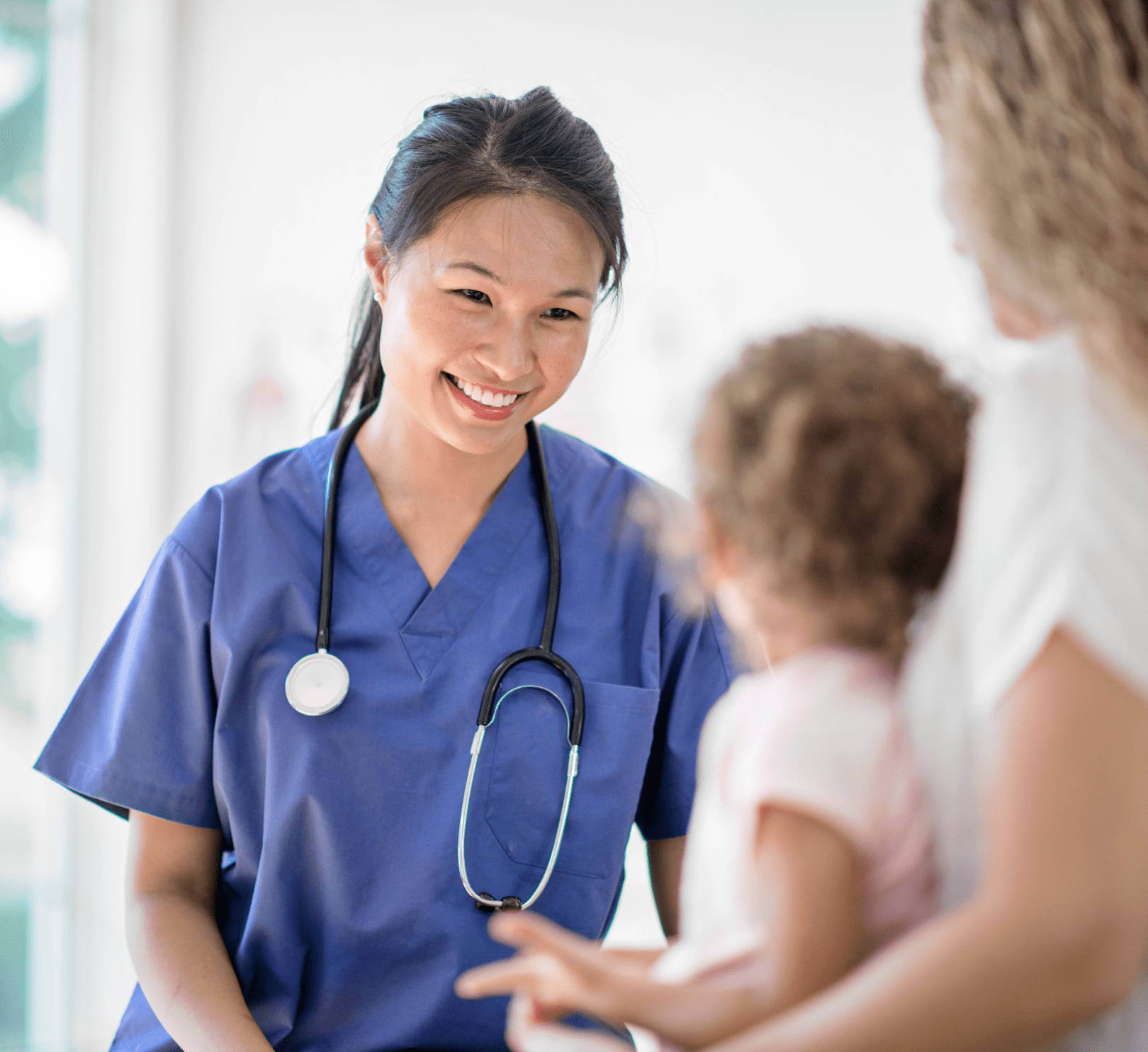 Une professionnelle de santé sourit alors qu’elle examine un jeune enfant assis sur les genoux de sa mère. Elle porte une blouse bleue et un stéthoscope autour du cou.