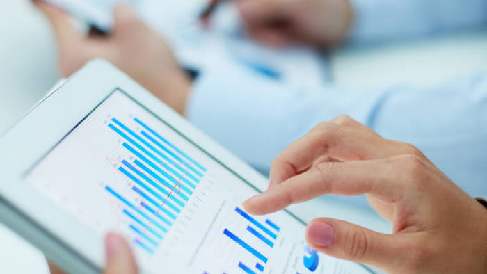 A close-up view of two people's hands operating tablet devices. One tablet is visible and shows a set of blue bar charts. The second tablet in the background is out of view.