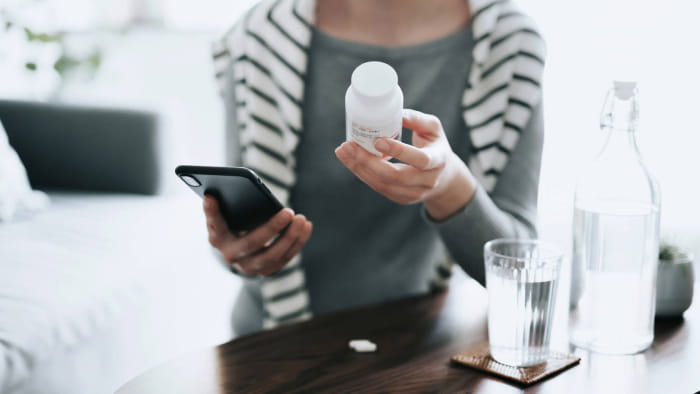 Une personne tenant une bouteille blanche de médicaments sur ordonnance dans une main et un téléphone cellulaire dans l’autre. Ils sont assis à une table avec un verre d’eau devant eux tout en portant une chemise grise à manches longues et un pull rayé noir et blanc.