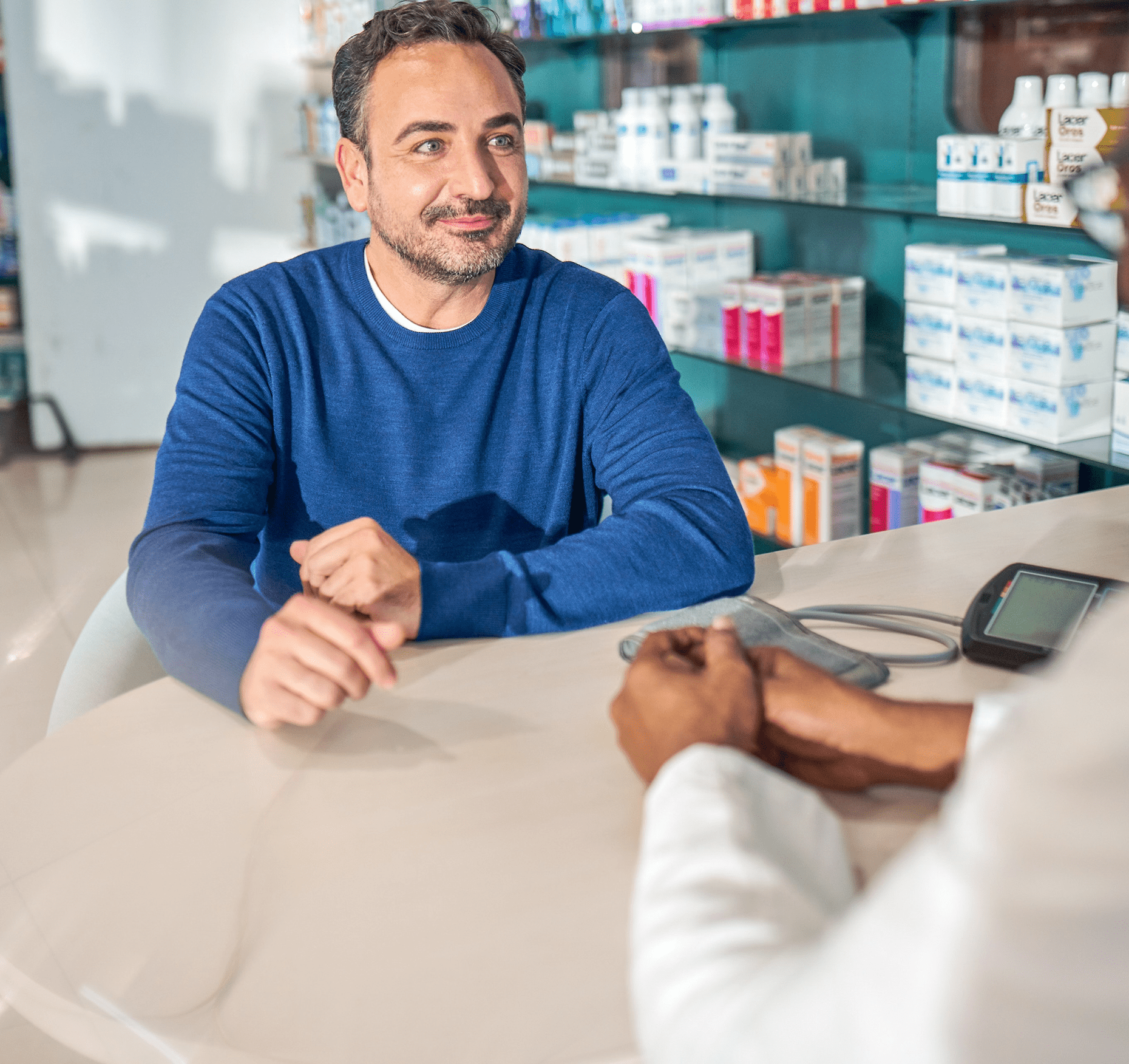 Man seated at a table inside a pharmacy, consulting with a pharmacy staff member, with a blood pressure monitor on the table.