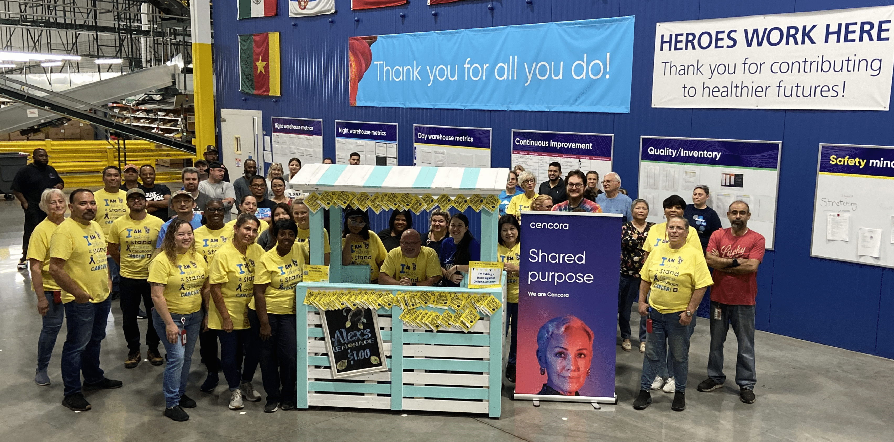 A large group of volunteers pose for a photo in a Cencora warehouse, surrounding a blue and white-striped lemonade stand. 