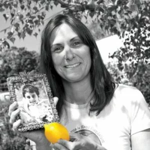 Photograph of Liz Scott, Alex’s mom and Co-Executive Director of Alex’s Lemonade Stand Foundation. Liz holds a framed photo of a child in one hand and a lemon in the other. The photo is black and white, except for the yellow lemon, and is taken outside under a tree.