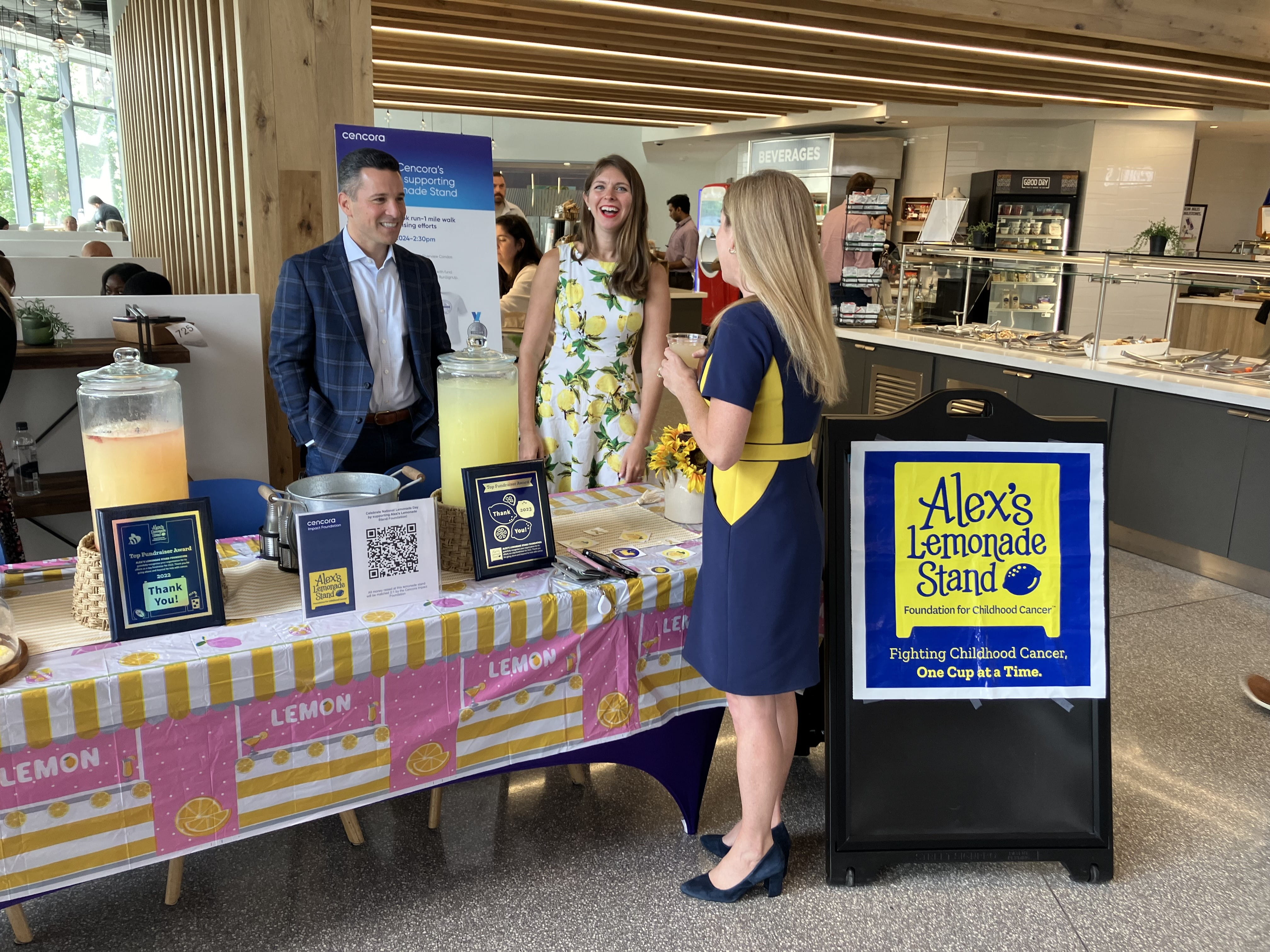Two people stand behind a table in the cafeteria at Cencora’s office. The table displays signage for Alex’s Lemonade Stand next to two large jugs of lemonade. Someone stands on the other side of the table and holds a cup of lemonade. A large plastic signboard says, “Alex’s Lemonade Stand. Fighting childhood cancer. One cup at a time.”