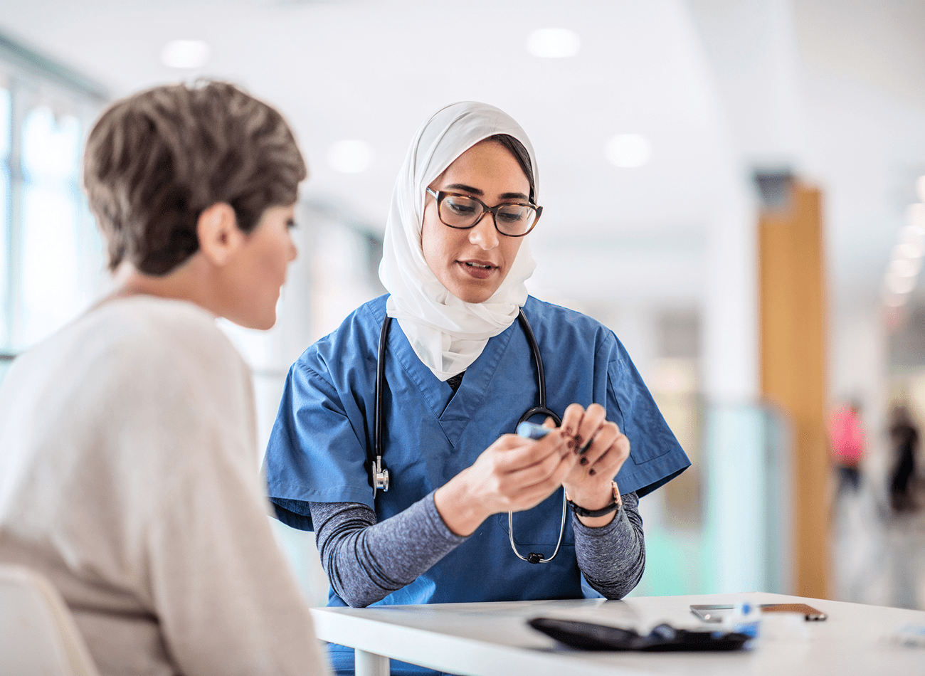A medium-light-skinned woman wearing dark eyeglasses and a white hijab shows a light-skinned woman wearing a light brown sweater a vial of medicine.