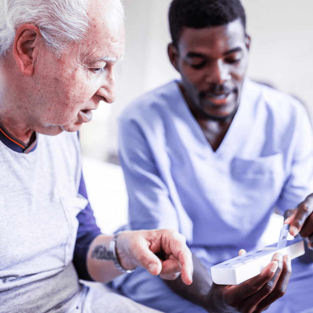 A young, dark-skinned person wearing blue scrubs talks to his elderly, light-skinned male patient about the medications in the pill box he holds in his hand.