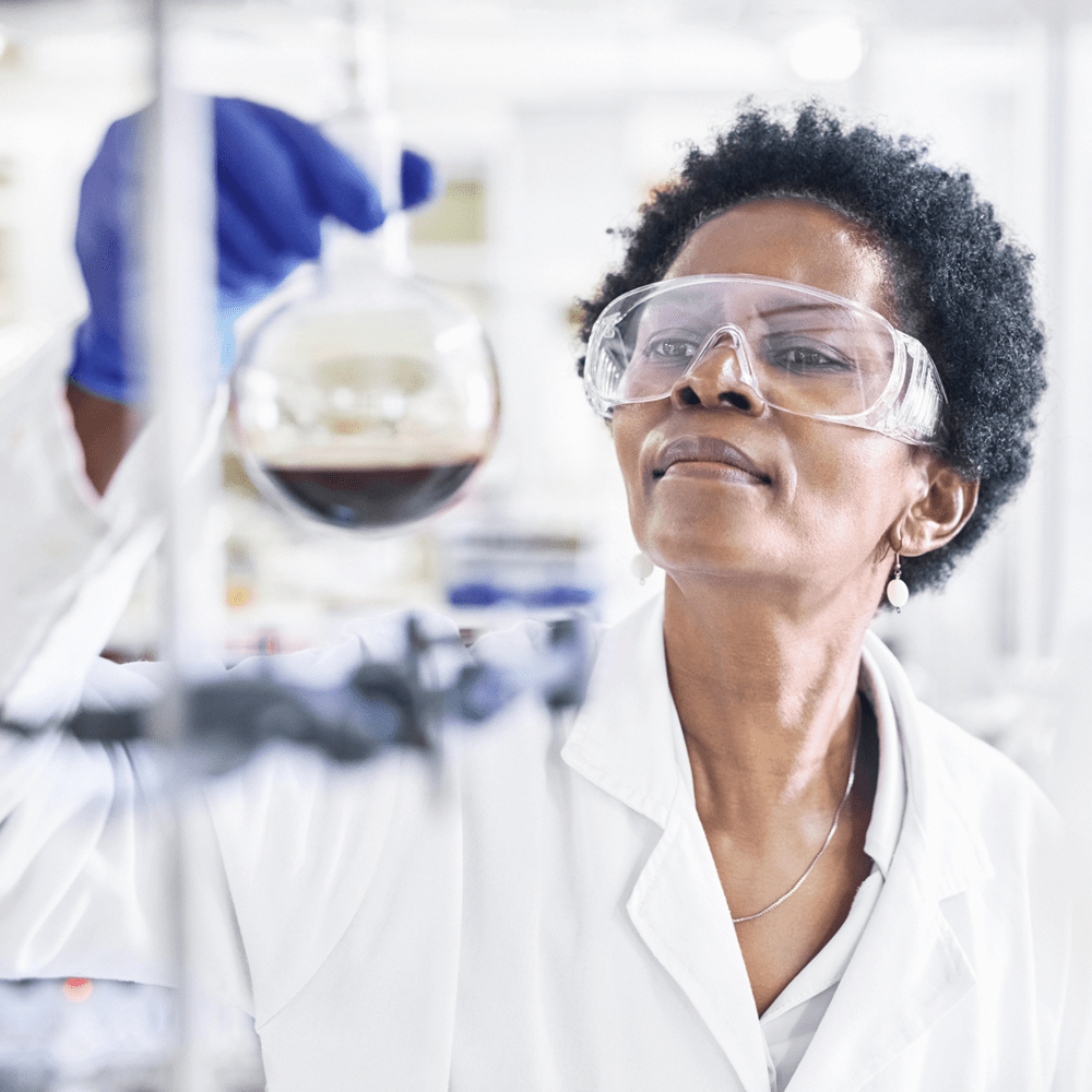 A dark-skinned person wearing a lab coat and goggles studies a dark solution in a glass container.